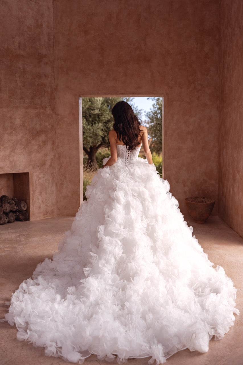 A woman viewed from behind wearing a voluminous white wedding gown with a heavily textured, ruffled tulle skirt, standing in a rustic room with textured brown walls looking out to a garden.