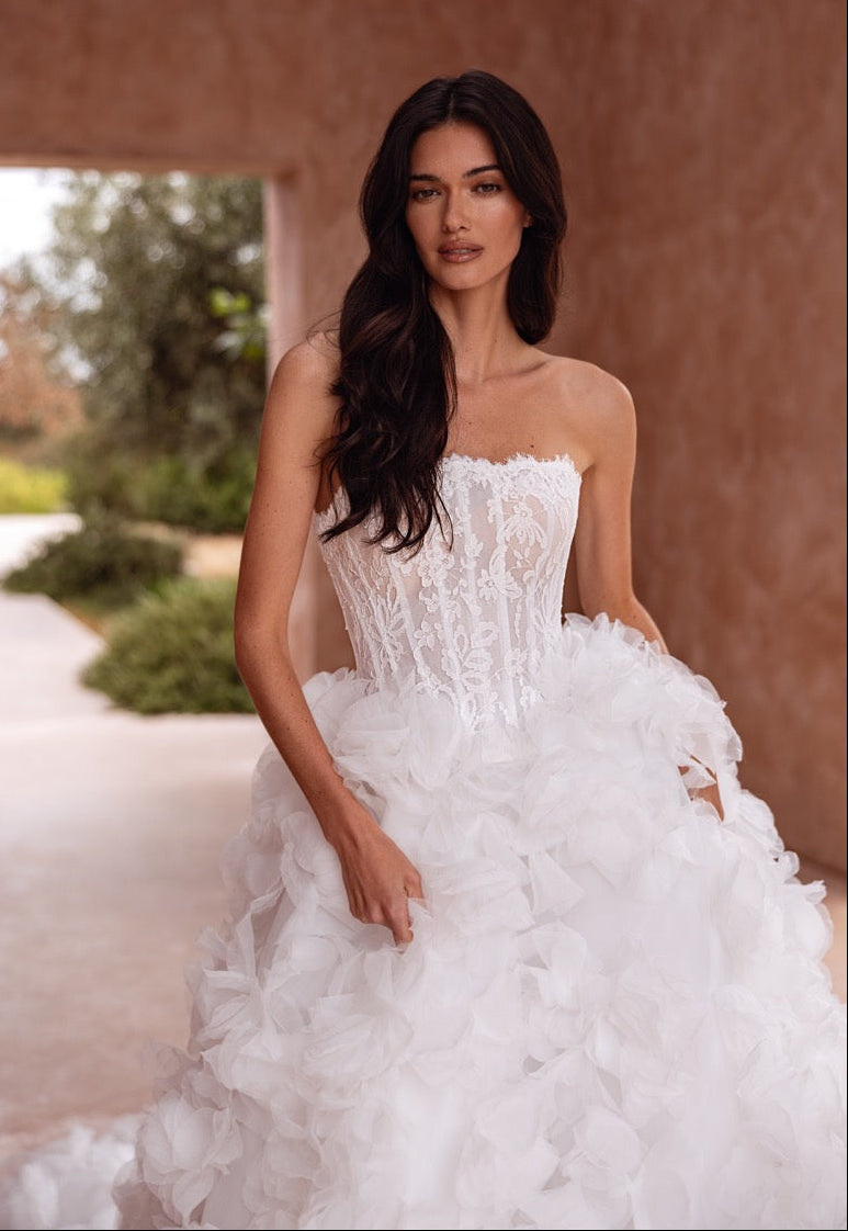 A woman wearing a strapless white wedding gown with a structured lace corset bodice and a voluminous ruffled tulle skirt, standing in a covered outdoor walkway with a warm stucco wall behind her.